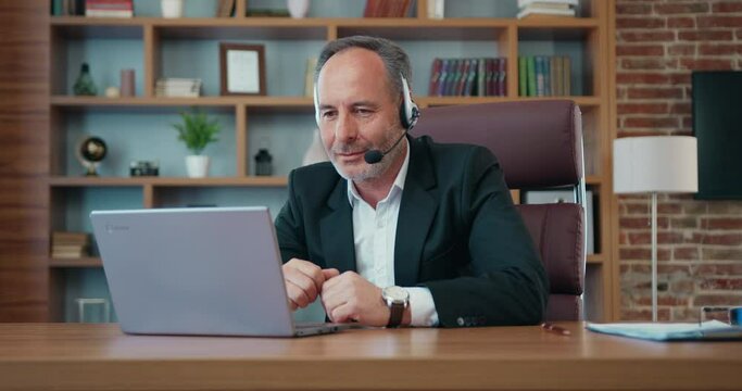 Attractive positive successful bearded businessman in headset holding videochat on computer with his business partners in modern office