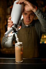 bartender holds siphon and decorates glass with cocktail with whipped cream