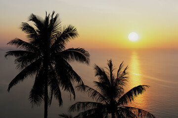 Drone aerial image view of silhouette coconut palm trees on the island with peaceful ocean, mountain and summer morning bright sunrise for travel and tourism industry background.