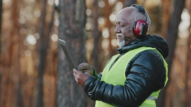 Elderly african american forestry engineer in noiseisolating headphones in forest old mature foreman forester thinks over work plan for cutting trees by entering data into tablet evaluating situation