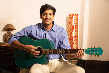 Obraz premium Happy Asian Indian young man playing acoustic guitar at home, music teacher or male musician.