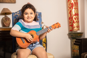 Happy little Indian girl playing acoustic guitar or ukulele at home, learning music.