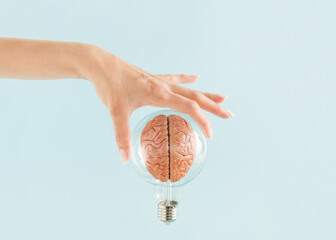 Female hand holds transparent light bulb with human brain on isolated pastel blue background....