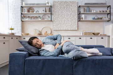 Sick and sad woman lying on the sofa at home in the living room, Asian woman has severe stomach...