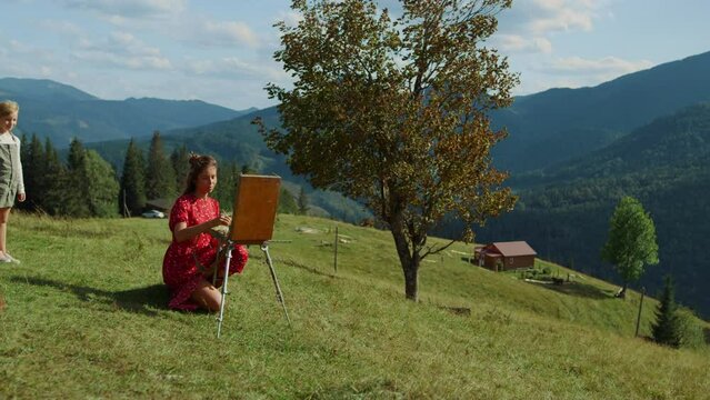 Creative woman drawing picture in mountains. Girl watching at painting process.