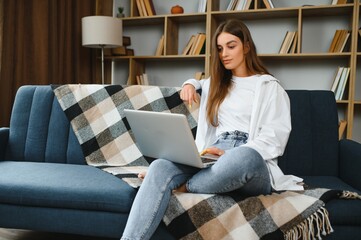 Smiling attractive young woman sitting on sofa using laptop communicating working online at home, happy teen girl typing on computer, enjoying writing blog or chatting with friends in social network