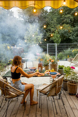 Woman cooks food on disposable grill while sitting relaxed by the table on cozy terrace during the evening at beautiful backyard