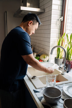 Adult brunette asian man wearing t-shirt washing dishes at kitchen
