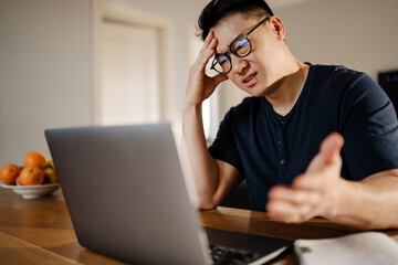 Adult tense asian man in glasses studying with laptop