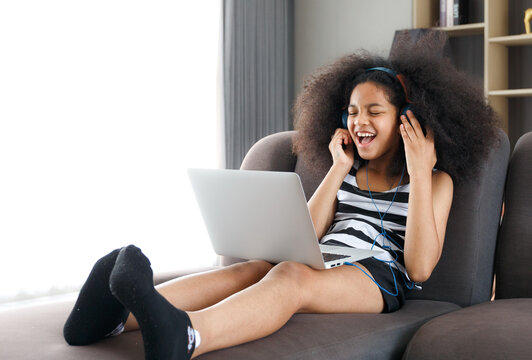 A cute little girl wearing headphones is sitting on her laptop on the sofa on her vacation.