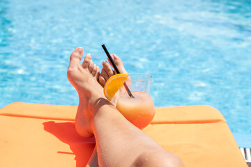 Fresh glass of a cocktail with an orange slice and woman legs on a sun lounger by a swimming pool. Holiday and vacation concept.