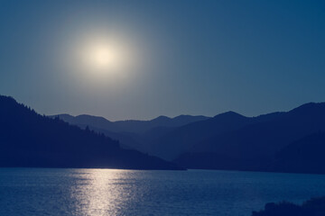 Mountain lake, the silhouette of the mountains and the surface of the water, the moon, the sun reflected in the water. Beautiful blue landscape