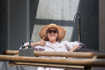 Front view medium shot portrait of a relaxed Asian woman wearing sunglasses, hat, and bathrobe,...