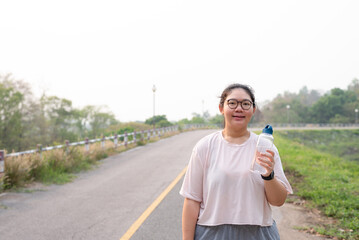 Front view, medium shot portrait with copy space of young fat Asian woman with glasses, holding water bottle at outdoor park in the morning, smiling, looking at camera. Sport and obesity concept.