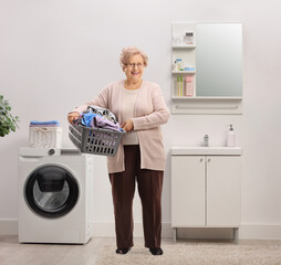 Full length portrait of an elderly woman holding a laundry basket with clothes in a bathroom - obrazy, fototapety, plakaty