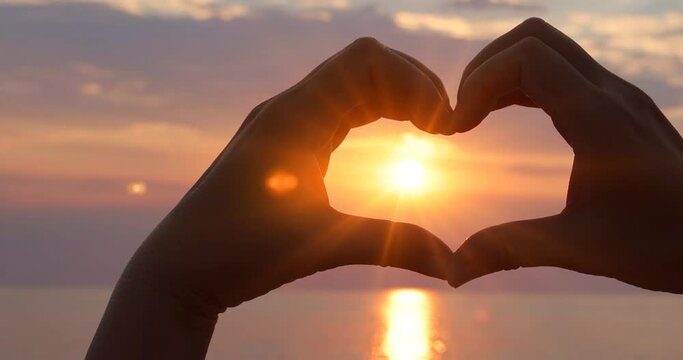Female hands in the form of heart with the sunlight passing through the hands at sunset on the sea. Hands in shape of love heart, slow motion. Love concept sea vacation