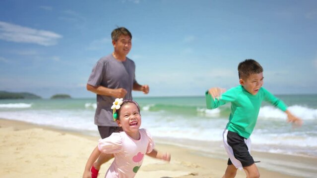 4K Happy Asian family on summer vacation. Grandfather jogging exercise with little grandchild boy and girl on the beach in sunny day. Senior man and children kid enjoy outdoor activity at the beach