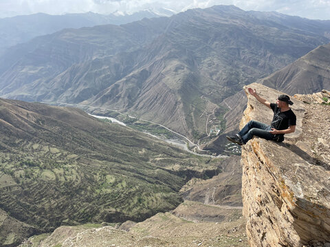 Male tourist admires mountains of North Caucasus while sits on edge of high mountain cliff, Goor village area in Dagestan