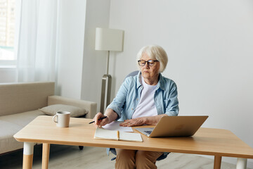 Fototapeta na wymiar A gray-haired, beautiful, stylish, serious lady works from home sitting at her desk with a laptop and notebooks for notes, in comfort