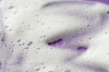 Foam swatch on a violet background. Soapy liquid texture with bubbles. Natural sunshine and shadows. Skin care cleansing cosmetic in top view.