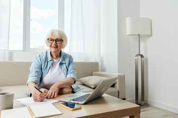 Handsome senior woman dressed in light blue shirt working with laptop at home sitting on sofa