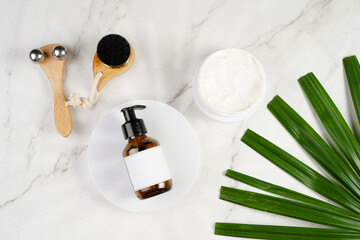 A mock-up of a brown cosmetics bottle with dispenser and white label on white round podium, round cream container, face brush, face massage roller on marble background and green palm leaf, top view
