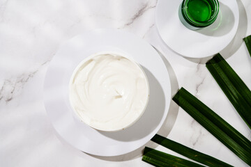 A mock-up of a round cosmetics container with cream on marble background and green palm leaf, top view