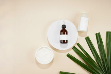 A mock-up of a brown bottle with pipette, white cosmetics container and container with cream on white round podium on beige colored background, green palm leaf,top view, space for text
