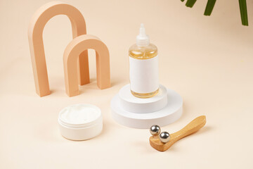 A mock-up of a transparent cosmetics bottle with yellow liquid and white label on white round podiums, container with white cream, face massage roller on beige colored background