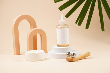 A mock-up of a transparent cosmetics bottle with yellow liquid and white label on white round podiums, container with white cream, face massage roller on beige colored background