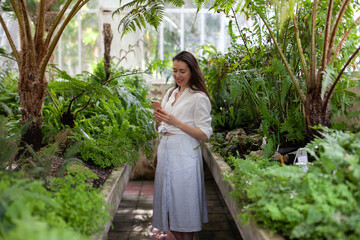 Obraz premium Young woman using mobile phone in greenhouse. Female tourist taking selfies or video chatting in nature.