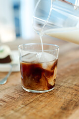 Pouring milk into a glass of black iced coffee.