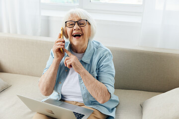 portrait of a happy elderly woman laughing happily during a phone conversation, pointing at him with her finger while sitting on the sofa in a bright apartment holding a laptop on her lap