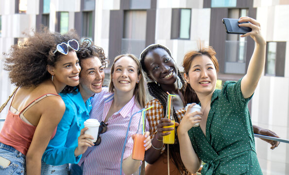 Multiracial group of happy 5 girls taking selfie on smartphone while enjoying sunny day in the city center . Different young millennial five friends take pictures with phone in hand with drink
