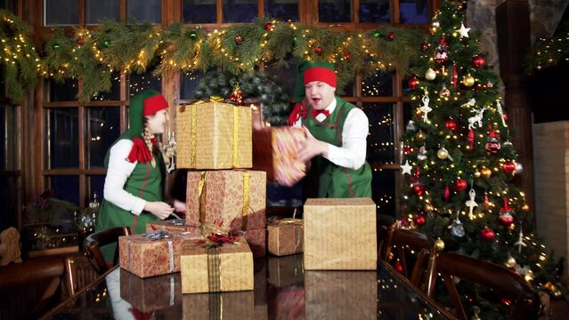 Presents for xmas holidays. Cheerful elves putting gift boxes on the table near Christmas tree in decorated room.