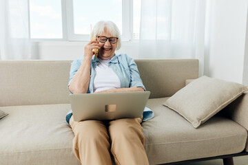 laughing loudly during a phone conversation with her family, an elderly lady is sitting relaxed on the couch holding a laptop on her lap while working remotely