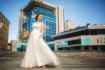 A luxurious girl in a wedding dress stands against the backdrop of a modern building with panoramic mirrored windows.
