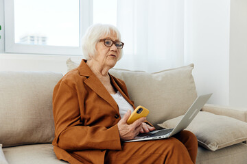 a sad, thoughtful elderly woman with gray hair is sitting on a cozy sofa dressed in a stylish brown suit and holding a laptop and smartphone sadly looking at the monitor