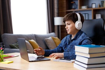 Schoolboy wearing earphones has online lesson, video call with teacher, answering questions, check of knowledge. Cute teenager studying at home using laptop. Distance learning and home education