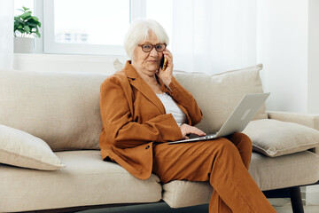 a lovely elderly woman is sitting on a wide beige sofa talking on a smartphone holding a laptop on her lap in a stylish brown suit