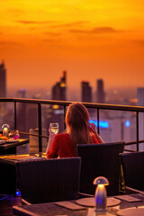 Young woman with glass of wine rest at luxury rooftop restaurant watching orange sky sunset. Female...