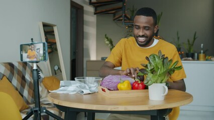 Creative man recording video for culinary vlog showing vegetables with high-five gesture using modern smartphone camera at home in kitchen