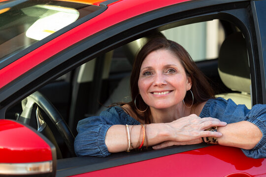 Beautiful middle aged brunette woman with brown eyes smiling in her red car while looking out the window at the viewer