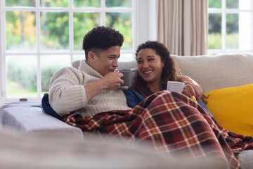Happy biracial couple sitting on couch under blanket, drinking coffee and laughing in living room