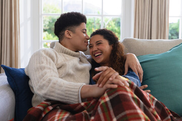 Happy biracial couple sitting on couch under blanket, laughing and embracing in living room - Powered by Adobe