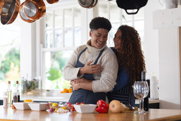 Happy biracial couple preparing food, smiling and embracing in kitchen