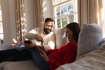 Happy caucasian couple sitting on couch in living room laughing, using smartphone and tablet