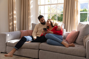 Happy caucasian couple sitting on couch in living room, talking and embracing, looking at smartphone
