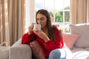 Happy caucasian woman sitting on couch drinking coffee in sunny living room