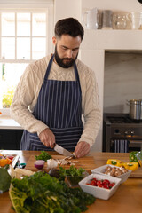 Caucasian man preparing food, concentrating on chopping vegetables, wearing apron in kitchen